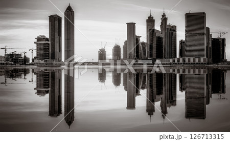 Monochrome city skyline with tall skyscrapers reflecting on calm water under a cloudy sky, Dubai Marina bay UAE 126713315