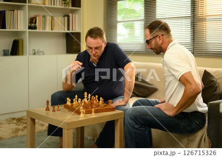 Father and adult son enjoying a game of chess in cozy living room. Fathers day concept. Father and adult son enjoying a game of chess in cozy living room. Fathers day concept. 126715326