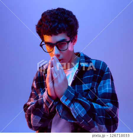 Teen boy in glasses holding hands in hopeful gesture, showing focused and serious mood in neon light against gradient background Teen boy in glasses holding hands in hopeful gesture, showing focused and serious mood in neon light against gradient background 126718033