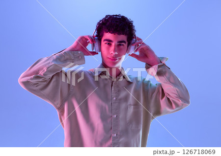 Teen boy, dressed casualwear, puts on headphones with slight smile, enjoying sound of musical immersion in neon light against gradient background Teen boy, dressed casualwear, puts on headphones with slight smile, enjoying sound of musical immersion in neon light against gradient background 126718069