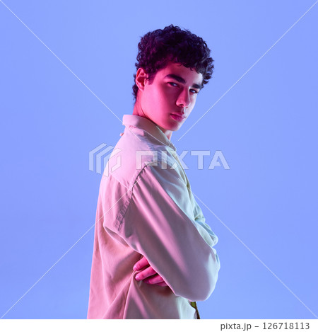 Stylish young man posing sideways with arms crossed on chest, with serious facial expression in contrasting neon lights against gradient background. Stylish young man posing sideways with arms crossed on chest, with serious facial expression in contrasting neon lights against gradient background. 126718113