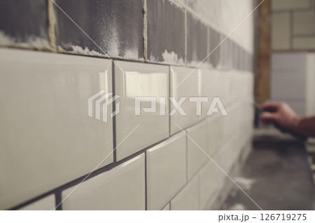 Close-up of a male hand applying grout on glossy ceramic wall tiles. Modern white, gray, and black subway-style tiles for interior renovation. Concept of home improvement and tiling work. Banner 126719275