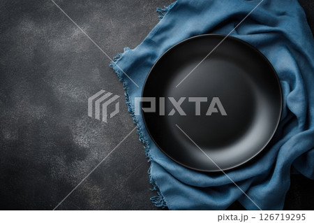 Empty matte black plate on turquoise fabric napkin over dark textured surface. Minimal top-down composition perfect for menu mockups, food blogs, restaurants, or elegant culinary presentations 126719295
