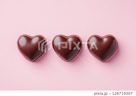 Top view of three heart-shaped dark chocolate candies placed in a row on soft pink background. Minimal design. Sweet romantic mood for Valentine's Day, gifts or dessert branding. Copy space. Banner. 126719307