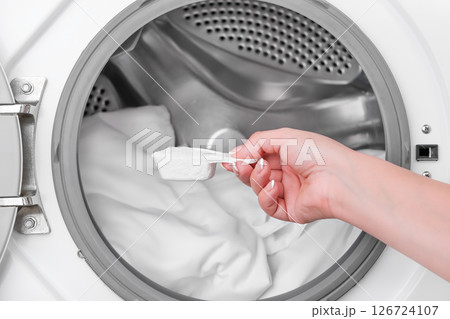 hand holding measuring spoon with washing powder against background of washing machine with white laundry hand holding measuring spoon with washing powder against background of washing machine with white laundry 126724107