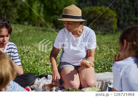 A family gathers for a joyful summer picnic in a scenic mountain meadow. The mother delights in a taco while surrounded by children, embracing the beauty of nature and family bonds. A family gathers for a joyful summer picnic in a scenic mountain meadow. The mother delights in a taco while surrounded by children, embracing the beauty of nature and family bonds. 126725968
