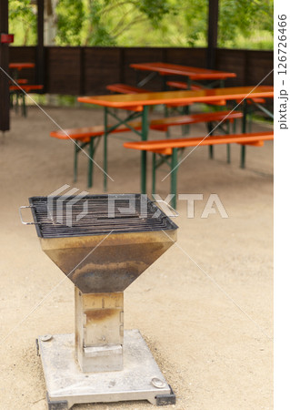 A charcoal BBQ grill stands in the center of a park's picnic area, colored orange tables surround it, creating a welcoming spot for gatherings amidst lush greenery 126726466