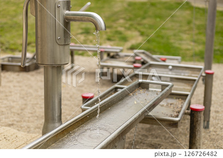A stainless steel water pump streams water into a channel at a playground, creating an engaging experience for children as they discover the fun of flowing water outdoors A stainless steel water pump streams water into a channel at a playground, creating an engaging experience for children as they discover the fun of flowing water outdoors 126726482