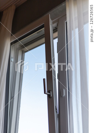 A white framed window is open, allowing sunlight to illuminate the room. Sheer curtains flutter gently, revealing a bright blue sky outside. The scene suggests a peaceful daytime atmosphere A white framed window is open, allowing sunlight to illuminate the room. Sheer curtains flutter gently, revealing a bright blue sky outside. The scene suggests a peaceful daytime atmosphere 126726565