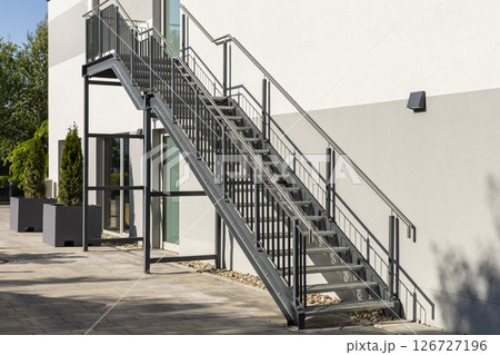 Sleek metal staircase connects the ground level to upper floor of contemporary building. Surrounding greenery adds touch of nature to the structured design. Metal construction, welding, steel stairs. 126727196