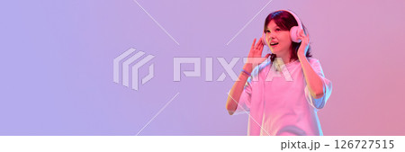 Teen girl with headphones joyfully talking waving hands showing excitement under vibrant light against gradient background with negative space. Banner 126727515