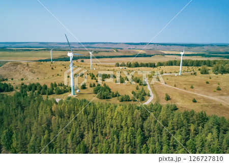 Tall wind turbines rise above rolling hills and a lush green forest, showcasing renewable energy efforts. Tall wind turbines rise above rolling hills and a lush green forest, showcasing renewable energy efforts. 126727810