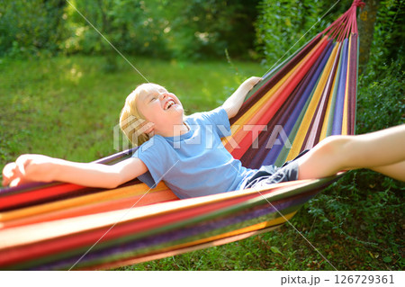 A cute blonde white preteen boy is having fun in the summer in a colorful hammock in the backyard or on the outdoor playground. A child is resting and swinging in a hammock. 126729361