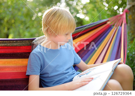 Cute preteen blond caucasian boy reading book and having fun with multicolored hammock in backyard or outdoor playground. 126729364
