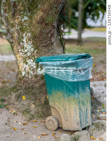 Trash bin with dirt in natural outdoor setting beside tree, showing urban waste and environmental decay 126730548
