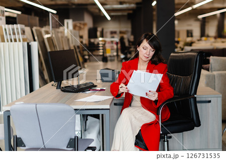 Saleswoman analyzing documents while sitting at desk in furniture store 126731535