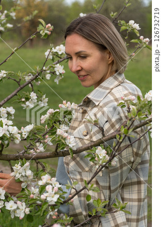 Woman in a plaid coat standing among blooming branches in a spring orchard, softly gazing through apple blossoms. Femininity, self-connection, beauty in nature, and spring renewal Woman in a plaid coat standing among blooming branches in a spring orchard, softly gazing through apple blossoms. Femininity, self-connection, beauty in nature, and spring renewal 126732719