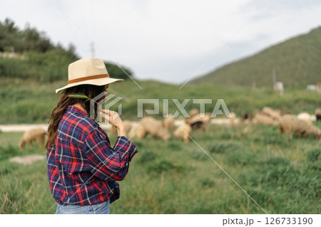 A woman wearing a straw hat stands in a field with a herd of sheep 126733190