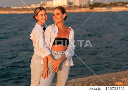 Couple Sunset Beach Vacation: Two women stand on a beach at sunset, facing the ocean, with a city skyline in the background. 126733202