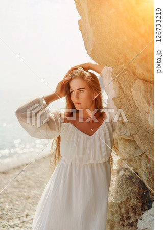 woman white dress standing on a beach, looking out at the ocean. She is lost in thought, with her hand on her hip. Concept of calm and introspection 126733219