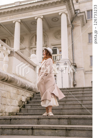 A woman in a hat stands on a set of stairs in front of a building. She is wearing a coat and a skirt. 126733233