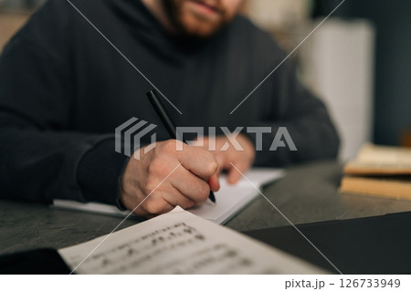 Close-up of music composer male writing melodic notes in home studio, sitting at table with laptop and sheet music scattered across workspace, merging artistic expression with digital tools Close-up of music composer male writing melodic notes in home studio, sitting at table with laptop and sheet music scattered across workspace, merging artistic expression with digital tools 126733949