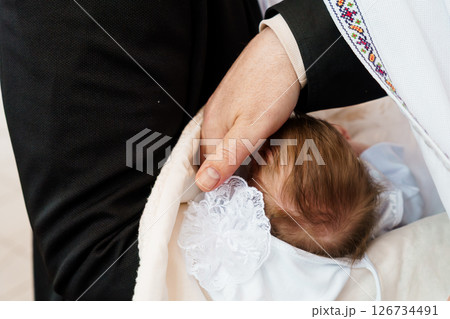 A tender moment of a baptism ceremony showcasing a man holding a baby wrapped in a white cloth with lace details, a priest's hand gently resting on the baby s head 126734491