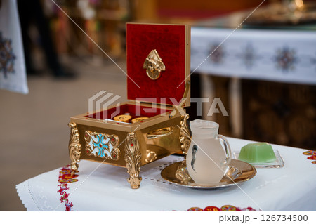 A richly ornamented red and gold reliquary box rests open on a white cloth alongside a glass of liquid and a small dessert, within a church setting 126734500