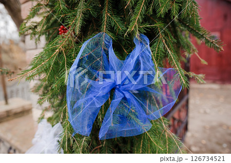 A vibrant blue tulle bow adorns a lush green evergreen wreath hung on a rustic red barn exterior, complemented by snow and decorative red berries, creating a festive winter scene A vibrant blue tulle bow adorns a lush green evergreen wreath hung on a rustic red barn exterior, complemented by snow and decorative red berries, creating a festive winter scene 126734521