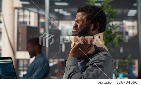 Hurt employee with a cervical collar working on a new business project, struggling to sit at the desk with his neck fracture. African american company associate tries to solve tasks. Camera A. 126734660