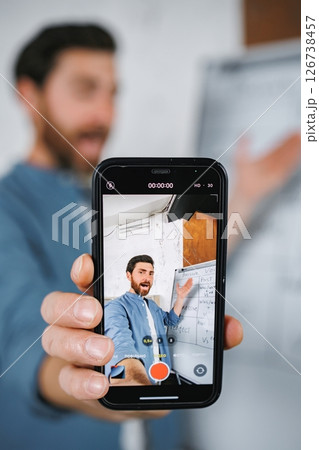 Cropped photo of a phone screen recording man explaining some language lesson. Male tutor sitting at table and recording a video of himself on a phone. Tutoring remote, modern education at home 126738457