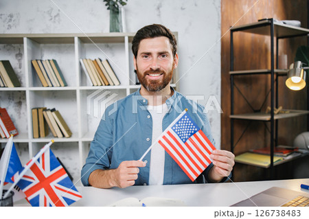 Excited male tutor sitting at table with flag of USA looking and smiling at camera, copy space. Man teaching foreign language online at home. Tutoring remote, modern education at home, online lesson Excited male tutor sitting at table with flag of USA looking and smiling at camera, copy space. Man teaching foreign language online at home. Tutoring remote, modern education at home, online lesson 126738483
