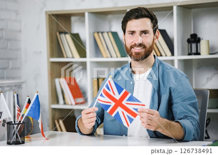 Excited male tutor sitting at table with flag of Great Britain, looking and smiling at camera, copy space. Man teaching foreign language online at home. Tutoring remote, modern education at home Excited male tutor sitting at table with flag of Great Britain, looking and smiling at camera, copy space. Man teaching foreign language online at home. Tutoring remote, modern education at home 126738491