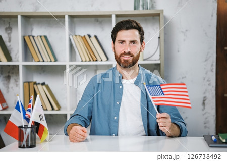 Excited male tutor sitting at table with flag of USA looking and smiling at camera, copy space. Man teaching foreign language online at home. Tutoring remote, modern education at home, online lesson Excited male tutor sitting at table with flag of USA looking and smiling at camera, copy space. Man teaching foreign language online at home. Tutoring remote, modern education at home, online lesson 126738492