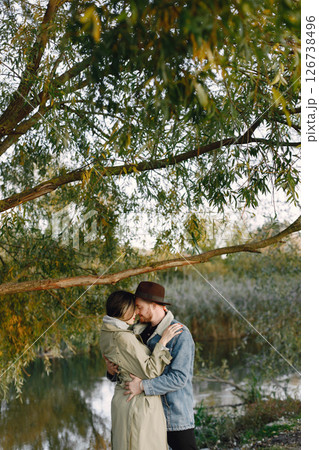 Man and woman in fashion clothes resting on a nature near the lake together. Man wearing jacket and a black hat and woman coat. Photo of romantic couple hugging. Man and woman in fashion clothes resting on a nature near the lake together. Man wearing jacket and a black hat and woman coat. Photo of romantic couple hugging. 126738496