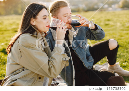 Romantic couple in fashion clothes sitting on a nature on a picnic rug. Man wearing jacket and woman coat. Woman and man drinking a wine. 126738502