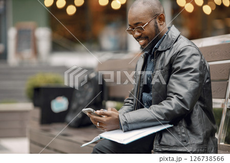 Black man sitting on a bench in a city. Man wearing sunglasses and a leather jacket. Business man has a papers and using a phone. 126738566
