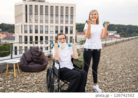Older couple engaging in light exercise with dumbbells, promoting recovery and health. Woman demonstrates activity while helping seated husband in wheelchair. Emphasizes fitness, rehabilitation Older couple engaging in light exercise with dumbbells, promoting recovery and health. Woman demonstrates activity while helping seated husband in wheelchair. Emphasizes fitness, rehabilitation 126742502