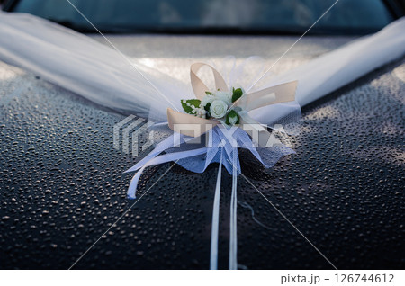Elegant wedding car decoration featuring white tulle draping over the hood with a delicate floral arrangement of roses and ribbons, wet surface reflecting light Elegant wedding car decoration featuring white tulle draping over the hood with a delicate floral arrangement of roses and ribbons, wet surface reflecting light 126744612