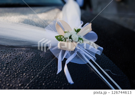 Wedding car decoration with a delicate white tulle bow and floral arrangement featuring roses, ribbons, and a heart-shaped detail black car roof 126744613