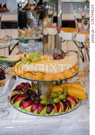 Exquisite Wedding Dessert Table with Tiered Fruit Display and Elegant Champagne Flutes, Featuring Apples, Bananas, Grapes, Cantaloupe and Pastries on Lace Cloth Exquisite Wedding Dessert Table with Tiered Fruit Display and Elegant Champagne Flutes, Featuring Apples, Bananas, Grapes, Cantaloupe and Pastries on Lace Cloth 126744624