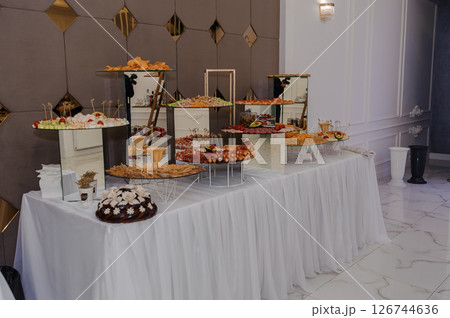 Elegant buffet table arrangement in a luxurious hall with white linens, tiered stands displaying various appetizers and desserts, ornate decor and a polished marble floor setting 126744636