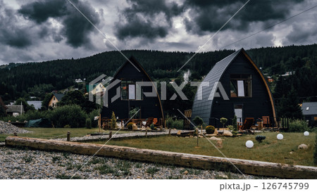 Two modern A-frame cabins sit side by side under a dramatic, stormy sky. Two modern A-frame cabins sit side by side under a dramatic, stormy sky. 126745799