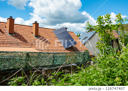 Solar panels installed on red-tiled roofs of houses surrounded by lush greenery and blue sky. Eco-friendly energy solutions in a rural setting highlight the blend of technology and nature. Solar panels installed on red-tiled roofs of houses surrounded by lush greenery and blue sky. Eco-friendly energy solutions in a rural setting highlight the blend of technology and nature. 126747407