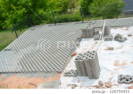 Concrete grid blocks are laid out at a construction site for a new driveway. Stacks of blocks and tools are visible, indicating ongoing work. Trees provide a lush background. 126750455