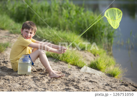 A little boy with a fishing net and a jar sits on the banks of a river or lake. 126751050