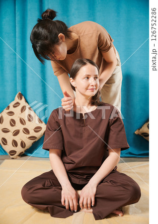 Masseuse making thai yoga massage. Woman therapist doing traditional massage treatment for female patient indoors. Client sitting on mat, meditating, enjoying hand massage in calm atmosphere. Masseuse making thai yoga massage. Woman therapist doing traditional massage treatment for female patient indoors. Client sitting on mat, meditating, enjoying hand massage in calm atmosphere. 126752286