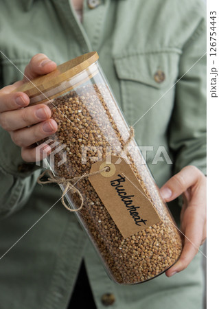 Unrecognizable woman holding refillable glass jar with raw buckwheat porridge. Refill product format eco-friendly living. Filling reusable container Whole spelt grain species. High protein fiber-rich Unrecognizable woman holding refillable glass jar with raw buckwheat porridge. Refill product format eco-friendly living. Filling reusable container Whole spelt grain species. High protein fiber-rich 126754443