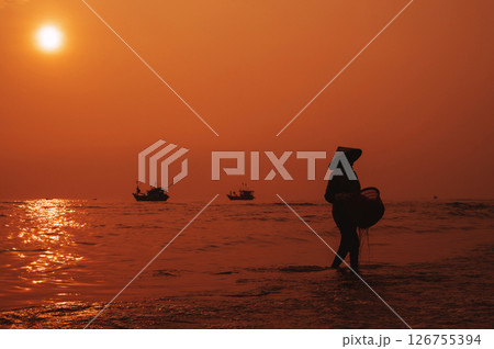 Silhouette of Vietnamese woman in straw hat on the shore of sea with ships in a fishing village in morning at sunrise in Vietnam 126755394