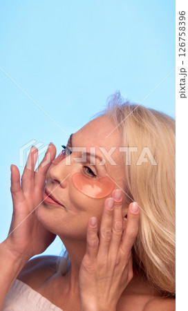 Senior woman gently adjusts peach-colored patches under her eyes, closing eyes and smiling softly while enjoying beauty routine. 126758396